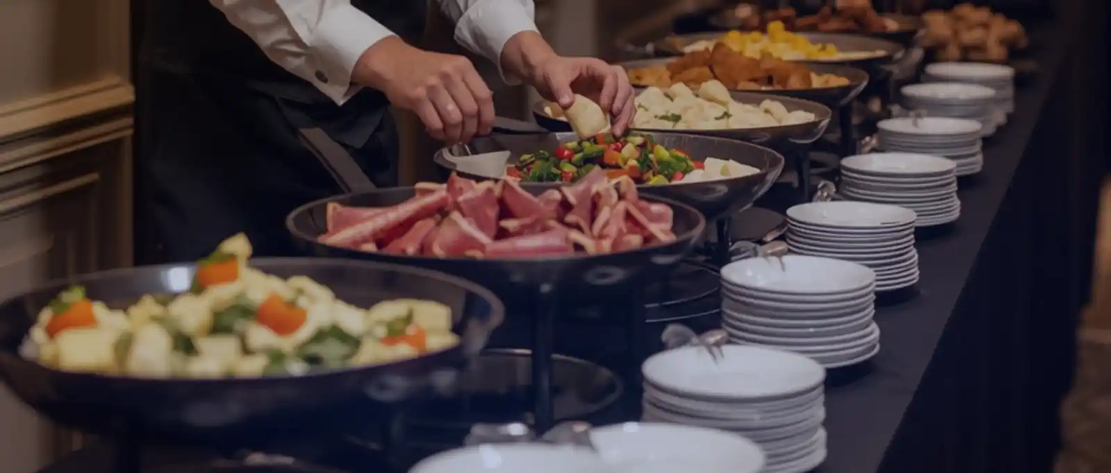 A professional caterer in uniform arranging fresh salads and side dishes on a buffet line next to stacks of clean white plates.