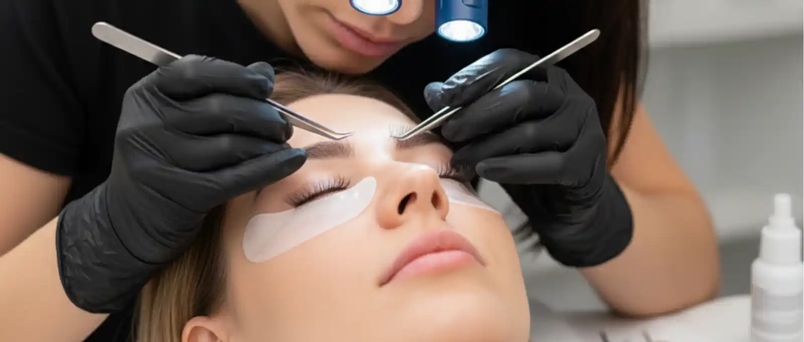 Lash technician performing eyelash extension treatment on client with professional lighting and protective eye pads